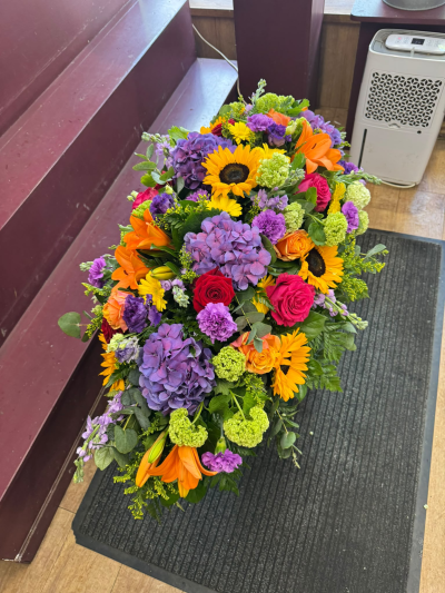A Colourful Celebration - A stunning, vibrant funeral spray bursting with beautiful shades of yellow, orange, pink and purple. Lovingly arranged with lush greenery, this colourful tribute is a wonderful way to celebrate a life filled with love, happiness and treasured memories. A heartfelt farewell to someone who brought colour and joy to the lives of others.
