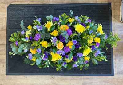 Sunshine and Serenity - A beautiful, full-length funeral spray featuring a cheerful combination of sunshine yellow, soft orange and rich purple tones, complemented by fresh, natural greenery. This elegant arrangement offers a lovely way to celebrate a life filled with warmth and happiness, creating a fitting and memorable final farewell.